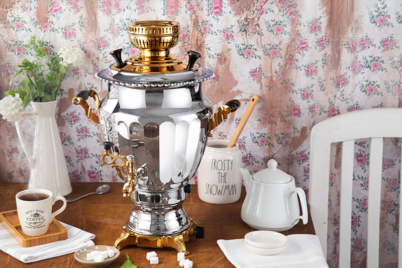 یک سماور گازی نقره‌ای و طلایی در کنار قوری و فنجان چای روی میز صبحانه